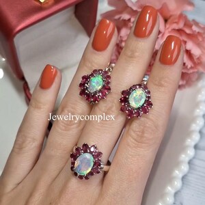 May include: A jewelry set featuring a ring and a pair of earrings. Each piece has a central oval opal surrounded by small, red gemstones. The jewelry is displayed on a hand with orange nail polish. The text "Jewelrycomplex" is visible.