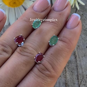 May include: Four oval-shaped stud earrings featuring two red rubies and two green emeralds set in silver prongs. The text Jewelrycomplex is visible in the centre. Each gemstone is individually mounted, showcasing a polished, faceted surface.