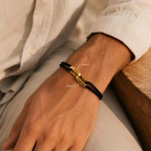 Può includere: Un bracciale in corda nera con due maglie metalliche rettangolari dorate intrecciate. Una maglia è incisa con il testo "PAPA" etichettato come Face 1, mentre l'altra maglia riporta il testo "D'AMOUR" etichettato come Face 2.