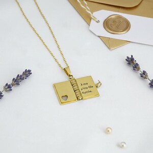Peut inclure: Un collier doré avec un médaillon en forme de livre. Le médaillon s'ouvre pour révéler un texte gravé. Une découpe en forme de cœur est visible sur le côté gauche. Le collier est sur une surface blanche, avec des brins de lavande et des perles.
