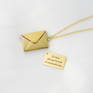 Peut inclure: Un collier doré avec un pendentif en forme d'enveloppe et une étiquette rectangulaire. L'étiquette porte l'inscription française "Je t'aime plus que les mots ne sauraient le dire." La chaîne est fine, sur fond blanc.