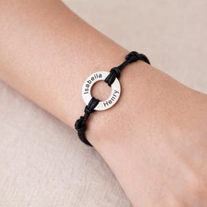 Peut inclure: Un bracelet en cordon noir avec un charm circulaire argenté. Le charm est gravé des noms "Isabella" et "Henry". Le bracelet est noué de chaque côté du charm.