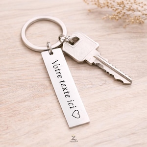 Peut inclure: Un porte-clés argenté avec une étiquette rectangulaire et une clé. L'étiquette porte le texte "Votre texte ici" et un symbole de cœur. Le porte-clés est attaché à un porte-clés et à une clé.