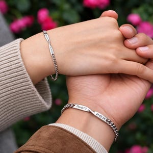 Op de afbeelding: Twee zilverkleurige kettingarmbanden met een centrale platte metalen staaf. De ene armband is gegraveerd met de naam Laura en de datum 29.02.24, de andere met de naam Daniel, een hartsymbool en de datum 29.02.24.