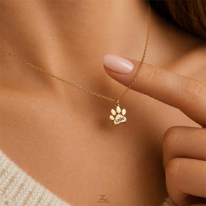 Pode incluir: Um colar de tom dourado com um pequeno pingente em forma de pata. O centro do pingente de pata está gravado com o nome Leiko em uma fonte clara e escura. O pingente está suspenso por uma corrente de metal fina e delicada.