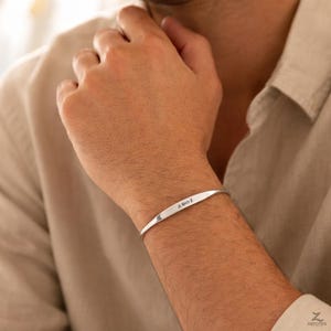 Peut inclure: Un bracelet manchette argenté avec texte gravé. Le bracelet est incurvé et porté au poignet d'une personne. Le texte gravé semble être des initiales et un symbole infini. La personne porte une chemise boutonnée de couleur claire.