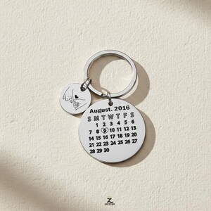 Peut inclure: Un porte-clés argenté avec deux breloques circulaires. L'une affiche le calendrier d'août 2016, avec un cœur le 9. L'autre montre deux mains entrelacées. Le porte-clés est posé sur une surface beige clair.