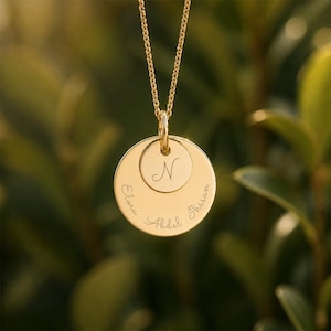 Peut inclure: Un collier pendentif doré avec une chaîne fine. Le pendentif comporte deux disques circulaires, l'un plus petit que l'autre. Le petit disque porte la lettre "N" gravée, et le grand disque porte les noms "Elora", "Abdel" et "Ihsan" gravés.