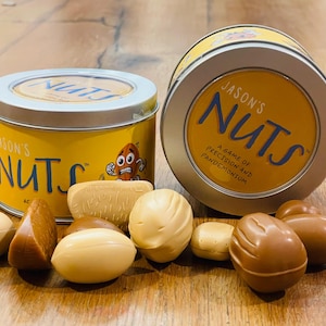以下が含まれることがあります： 「Jason's Nuts」の文字と漫画のナッツのキャラクターが描かれた、2つの黄色の金属缶。缶は開いており、茶色とタン色のナッツ型のゲームピースがいくつか表示されています。ゲームは「A Game of Precision and Pandemonium」です。
