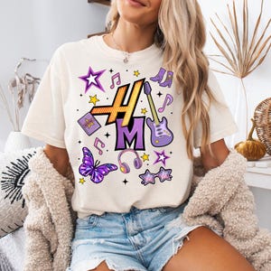 Peut inclure: T-shirt cr&egrave;me avec un motif graphique color&eacute;. Le motif comprend les lettres "H" et "M" en or et violet, ainsi que des notes de musique, des &eacute;toiles, un papillon, un casque et une guitare. Le t-shirt est un v&ecirc;tement d&eacute;contract&eacute; pour tous les jours.