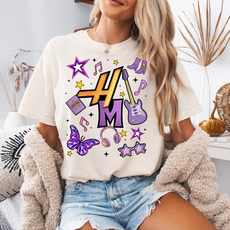 Peut inclure: T-shirt cr&egrave;me avec un motif graphique color&eacute;. Le motif comprend les lettres "H" et "M" en or et violet, ainsi que des notes de musique, des &eacute;toiles, un papillon, un casque et une guitare. Le t-shirt est un v&ecirc;tement d&eacute;contract&eacute; pour tous les jours.
