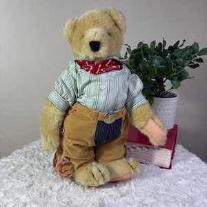 May include: A plush tan teddy bear wearing a light blue and white pinstriped button-down shirt, a red and white patterned bandana around its neck, and tan trousers with a dark blue panel, a silver-toned belt buckle, and decorative side ties.
