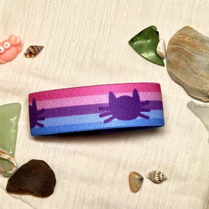 Puede incluir: Una pulsera de tela con un patrón de rayas horizontales en tonos rosa, morado y azul. La banda está decorada con una silueta repetida de color morado oscuro de una cara de gato con bigotes, centrada sobre el fondo de rayas.