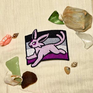 Puede incluir: Un parche bordado rectangular que presenta una criatura cuadrúpeda de color morado claro con orejas largas y cola, sobre un fondo de rayas horizontales negras, grises, blancas y moradas, rodeado de varias conchas pequeñas y cristales de mar.