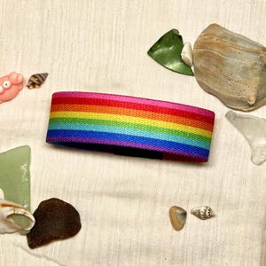 May include: A horizontal rainbow-striped elastic fabric band featuring vibrant red, orange, yellow, green, blue, and purple colors. The band is surrounded by various small sea shells, pieces of smooth sea glass, and a small pink crab figurine.