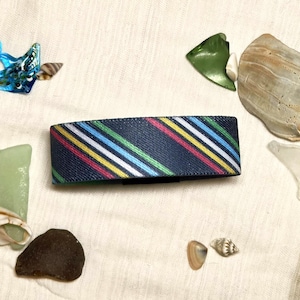 May include: A rectangular hair clip featuring a dark navy blue base with a series of diagonal stripes in vibrant shades of yellow, light blue, white, and pink. The accessory has a textured surface and is positioned horizontally in the centre.