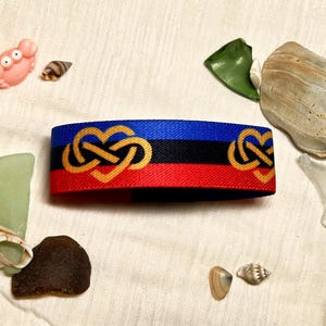 May include: A woven fabric wristband featuring horizontal stripes of blue, black, and red. The band is decorated with a repeating gold-colored infinity heart symbol pattern. The material has a textured, durable appearance with finished edges.