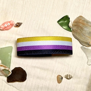 Può includere: Una fascia elastica rettangolare con quattro strisce orizzontali gialle, bianche, viola e nere. La fascia è circondata da varie piccole conchiglie, pezzi di vetro marino e una piccola statuetta di granchio rosa su tessuto bianco sporco.