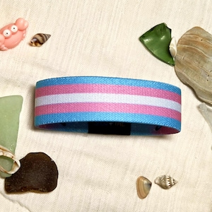 May include: A fabric wristband featuring horizontal stripes in light blue, pink, and white. The band is positioned centrally, surrounded by various small seashells, pieces of sea glass, and a small pink crab figurine on a textured fabric.