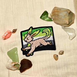 May include: A rectangular embroidered patch featuring a tan, fox-like creature with large ears and a leafy tail, set against a background of horizontal green, white, and black stripes. The patch has a thick black border and detailed stitching.