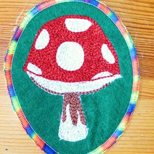 May include: An oval-shaped embroidered patch featuring a red mushroom with white spots on a dark green background. The patch is bordered by a vibrant, multi-colored rainbow thread edge that frames the central mushroom design clearly.