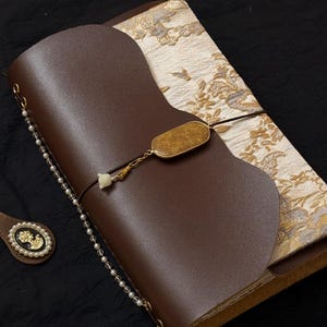 Pode incluir: Um diário de couro marrom com um fecho decorativo e uma corrente de contas. O diário tem um interior de tecido estampado e um pequeno enfeite redondo com um desenho floral.
