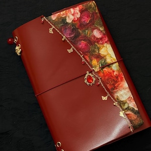 以下が含まれることがあります： 赤い革のジャーナル。表紙には花柄が施されています。蝶や赤い宝石など、チャームが付いたゴールドチェーンがジャーナルからぶら下がっています。ジャーナルは赤いゴムバンドで閉じられています。