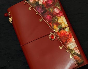 May include: A red leather journal with a floral patterned cover. A gold chain with charms, including butterflies and a red gem, dangles from the journal. The journal is closed with a red elastic band.