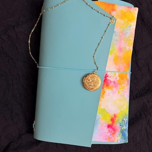 May include: A teal leather journal with a gold locket necklace attached. The journal is closed with a teal elastic band. The inside pages have a colorful watercolor design. The locket is round with a floral pattern.