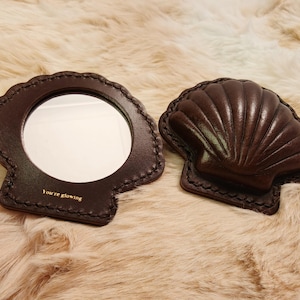 May include: Two brown leather shell-shaped compact mirrors. One mirror is open, revealing a circular mirror and the text "You're glowing". The other mirror is closed. Both have decorative stitching.
