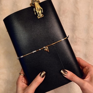 May include: Black leather journal with a gold-colored hand-shaped clip at the top. A gold beaded bracelet with a heart charm is wrapped around the journal. The journal is held by hands with gold-colored nail polish.