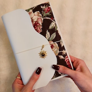 Könnte beinhalten: Ein weißes Notizbuch mit floralem Innenmuster und einem goldenen Sonnenanhänger. Das Notizbuch ist geöffnet und zeigt das florale Design. Die Hülle des Notizbuchs ist weiß und das florale Muster enthält rosa und cremefarbene Rosen.