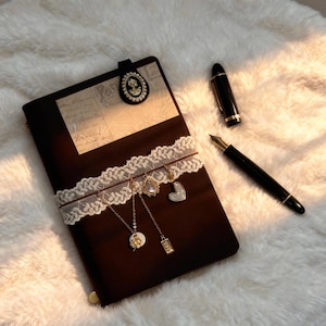 May include: A dark brown journal with a decorative white lace band and dangling charms. A black pen and a black and gold fountain pen are next to the journal. A cameo clip is attached to the top of the journal.