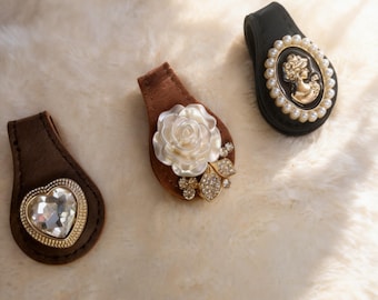 May include: Three decorative brown and black purse clips. One clip features a clear heart-shaped jewel, another has a white rose, and the third has a cameo surrounded by pearls. The clips are on a white, fluffy surface.