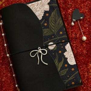 May include: Black leather journal with a floral and celestial patterned cover. A silver bow charm and a pearl chain dangle from the journal. A heart-shaped pin with pearl accents rests beside the journal on a red surface.