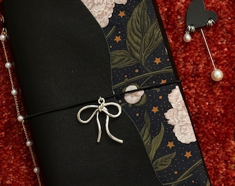 May include: Black leather journal with a floral and celestial patterned cover. A silver bow charm and a pearl chain dangle from the journal. A heart-shaped pin with pearl accents rests beside the journal on a red surface.