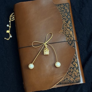 May include: A brown leather journal with a gold-colored charm and pearl accents. The journal has a decorative gold pattern on the corner and is secured with a black elastic band. A gold chain with pearls and a charm hangs from the top.