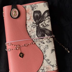 Puede incluir: Un diario de cuero rosa con una cubierta estampada floral y un cierre de cuentas decorativo. El diario presenta un broche negro con un emblema dorado y un dije de cuentas. La cubierta tiene un diseño curvo.