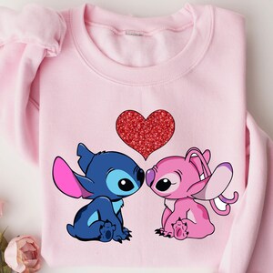 A pink sweatshirt with a graphic of two cartoon characters, a blue one and a pink one, sitting next to each other and a heart in the center.