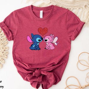 A red t-shirt with a graphic of two cartoon characters, a blue one and a pink one, standing close together and holding a heart.