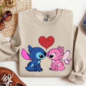 A beige sweatshirt with a graphic of two cartoon characters, a blue one and a pink one, sitting next to each other and a red heart in the center. The sweatshirt is placed on a white surface, along with a pair of sunglasses, a pair of jeans, and a pair of brown shoes.