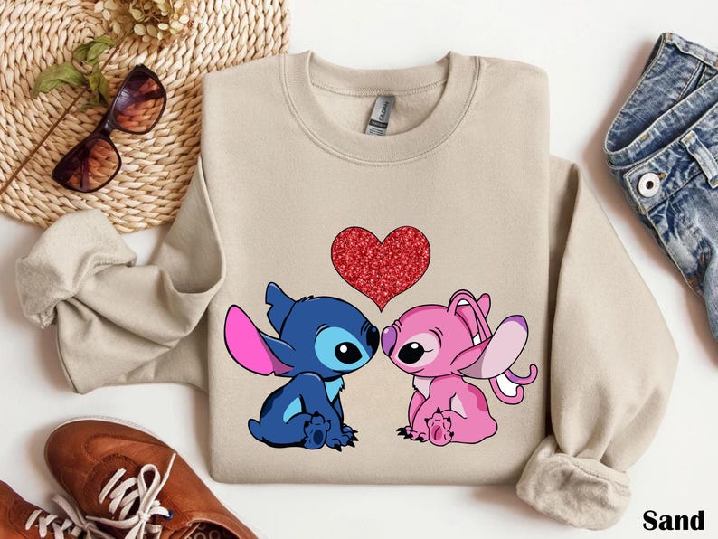 A beige sweatshirt with a graphic of two cartoon characters, a blue one and a pink one, sitting next to each other and a red heart in the center. The sweatshirt is placed on a white surface, along with a pair of sunglasses, a pair of jeans, and a pair of brown shoes.