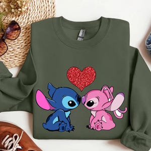 a green sweatshirt with a graphic of two cartoon characters, a blue character and a pink character, sitting next to each other and looking at each other. There is a red heart in the center of the image.