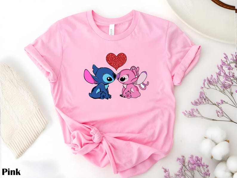 A pink t-shirt with a graphic of two cartoon characters, a blue one and a pink one, with a heart between them.