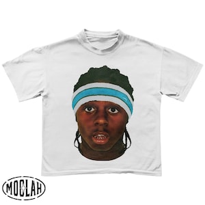 Puede incluir: Camiseta blanca con un retrato de una persona que lleva una cinta para la cabeza azul y blanca. La persona tiene la piel oscura y mira directamente al espectador. La camiseta tiene la palabra "MOBLAH" impresa en la parte inferior.