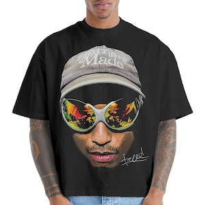 Puede incluir: Camiseta negra con un gráfico de una persona con gafas de sol y una gorra con el texto "Human Made". Las gafas de sol reflejan una escena colorida. La palabra "freeee!" está escrita en cursiva.