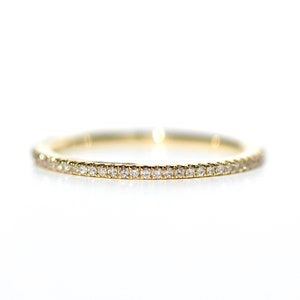 Puede incluir: Un delicado anillo de banda de oro, completamente engastado con peque&ntilde;os diamantes brillantes. El anillo se presenta sobre un fondo blanco brillante, resaltando los intrincados detalles y el brillo de las piedras preciosas.