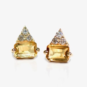 May include: A pair of gold-toned earrings. Each earring features a rectangular, yellow gemstone below a triangular arrangement of smaller, clear gemstones. Small, round accents are on either side of the rectangular stone.