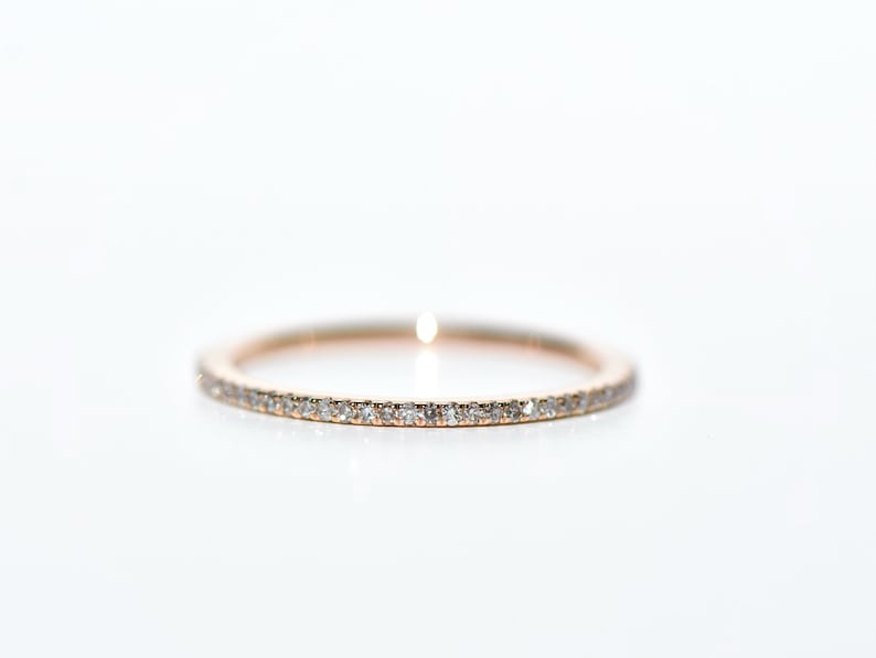 Puede incluir: Un anillo de banda de oro rosa con peque&ntilde;as piedras preciosas transparentes engastadas alrededor de toda la circunferencia. El anillo est&aacute; sobre un fondo blanco brillante, resaltando el brillo de las piedras preciosas. Este anillo es una joya fina.
