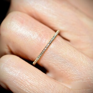 Puede incluir: Primer plano de un anillo de banda de oro engastado con peque&ntilde;as piedras preciosas transparentes. El anillo es delgado y delicado, con las piedras preciosas muy juntas alrededor de la banda. El anillo est&aacute; en el dedo de una persona.